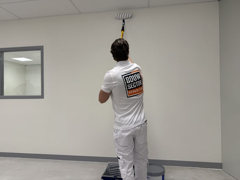 Schilder in witte werkkleding rolt een lichte muur tot aan het plafond, met verfbak en emmer op de vloer