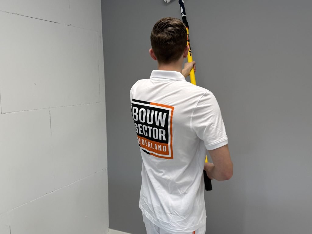 Schilder in wit poloshirt met Bouwsector-logo rolt een grijze wand naast een witte blokkenmuur