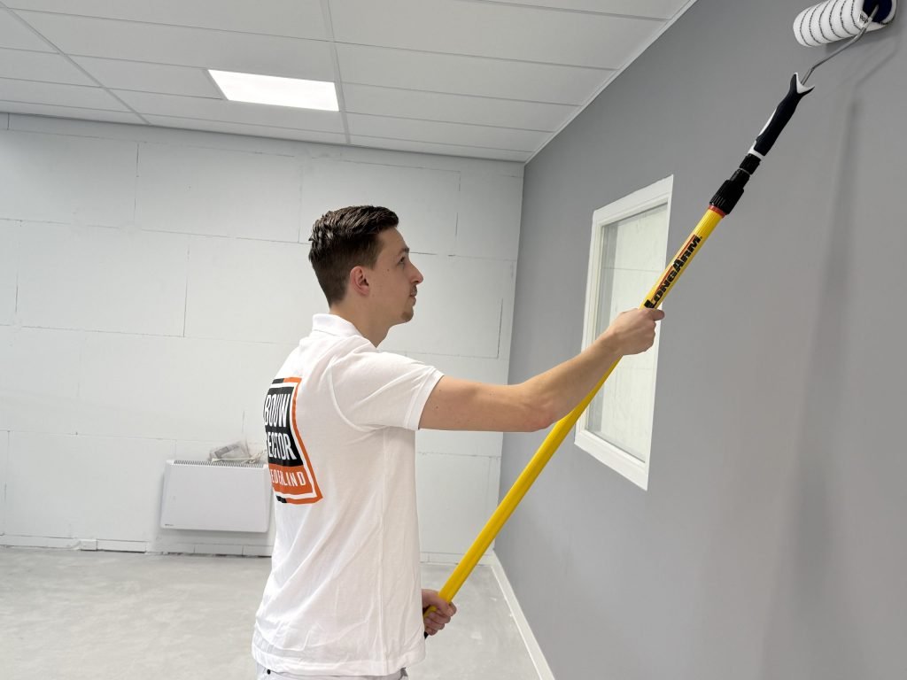 Schilder rolt een grijze wand met een telescoopsteel in een ruimte met een wit plafond van platen