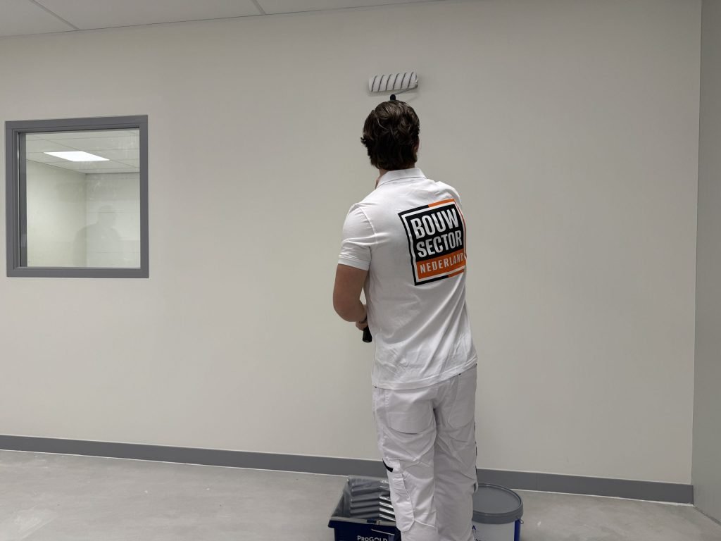 Schilder van Bouwsector Nederland rolt een witte wand in een lege ruimte, met verfbak en emmer aan zijn voeten